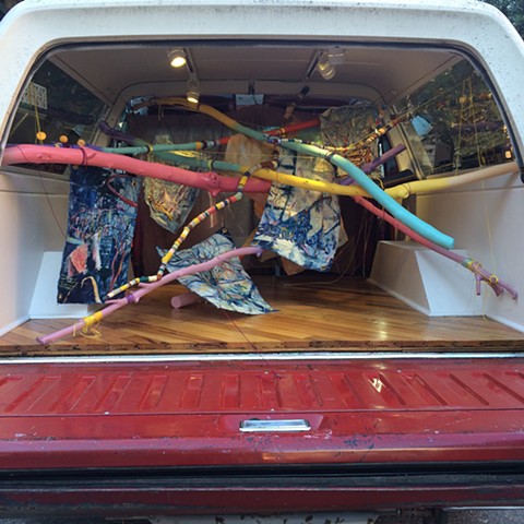 Brightly painted sticks crossing each other, providing hanging system for unstretched oil paintings on indigo dyed canvas, all installed in the back space of a 1991 Ford Bronco