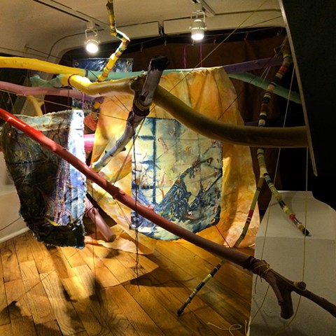Brightly painted sticks crossing each other, providing hanging system for unstretched oil paintings on indigo dyed canvas, all installed in the back space of a 1991 Ford Bronco