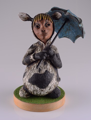 Girl seated and dressed in a Friesian cow onesie. She holds a blue umbrella and looks upwards.